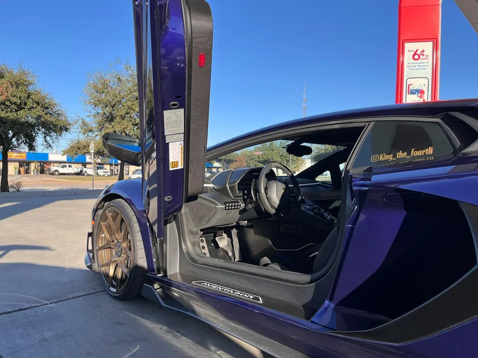 A purple Lamborghini Aventador with custom bronze wheels and open scissor doors at a gas station.