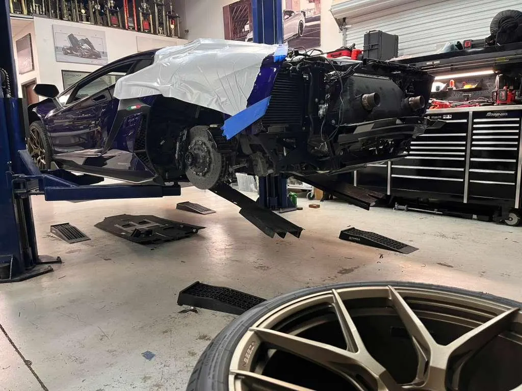 A purple Lamborghini Aventador on a lift in a performance shop undergoing disassembly, with a bronze wheel in the foreground.