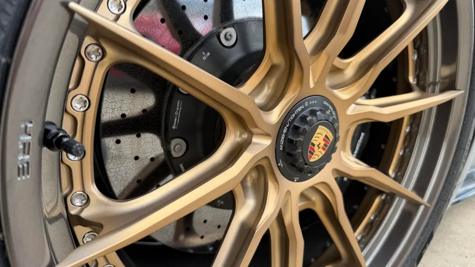 A close-up of a gold HRE wheel with a Porsche center cap and carbon ceramic brakes.