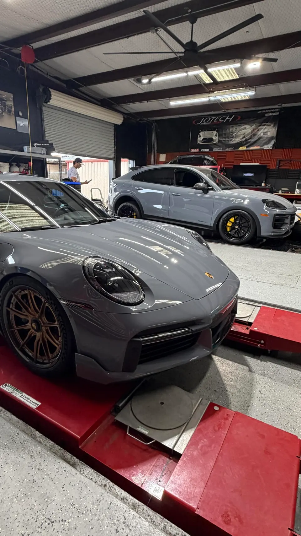 Two grey Porsche cars, a 911 and a Cayenne Coupe, inside a performance tuning shop with a person in the background.