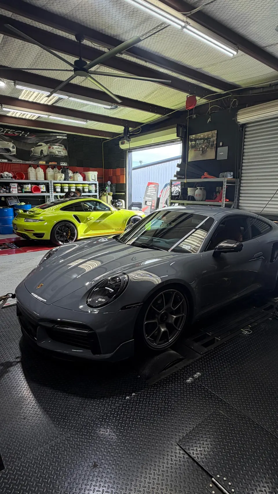 Two Porsche 911s, a grey 992 on a dyno and a yellow 992 parked behind it, inside a performance tuning shop.
