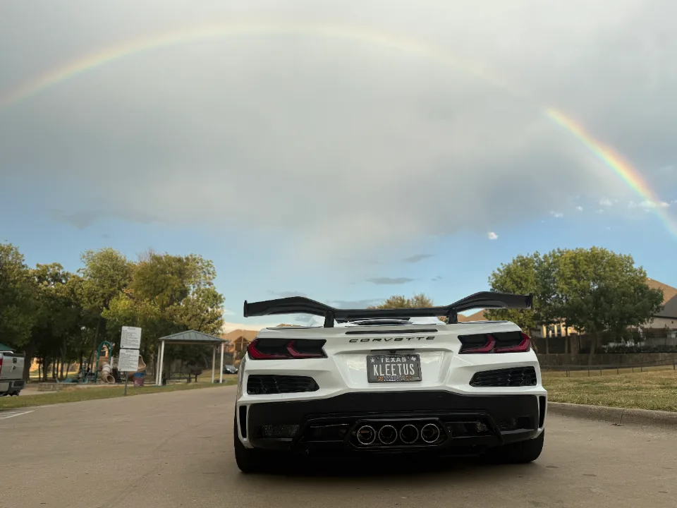 A white C8 Corvette with a large aftermarket rear wing parked outdoors under a vibrant rainbow.