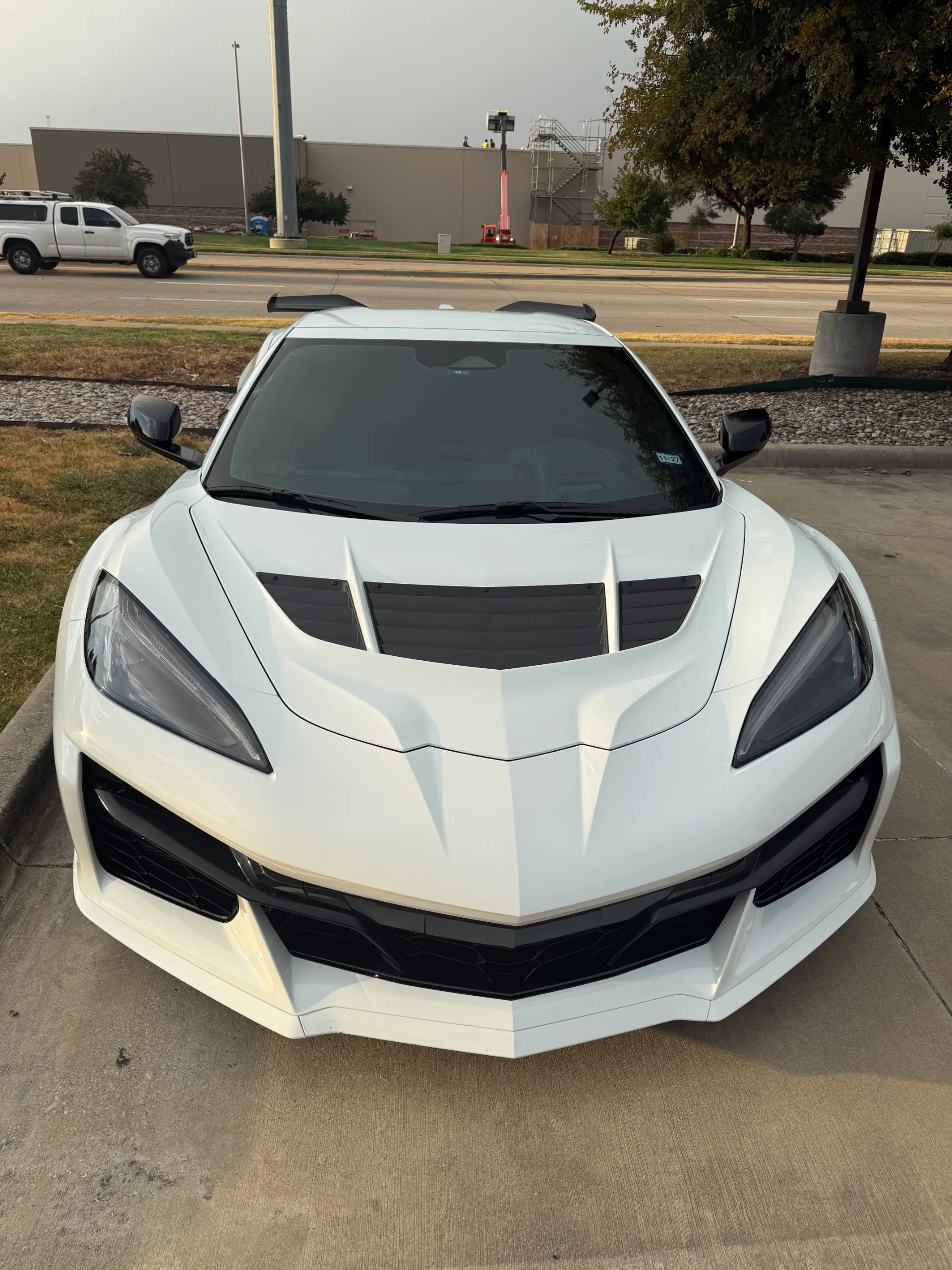 Front view of a white C8 Corvette parked on concrete with a street and buildings in the background.