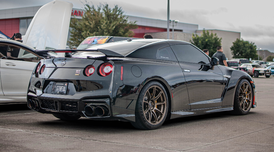 Rear three-quarter view of a black Nissan GT-R R35 with custom bronze wheels and aftermarket exhaust at an outdoor car event.