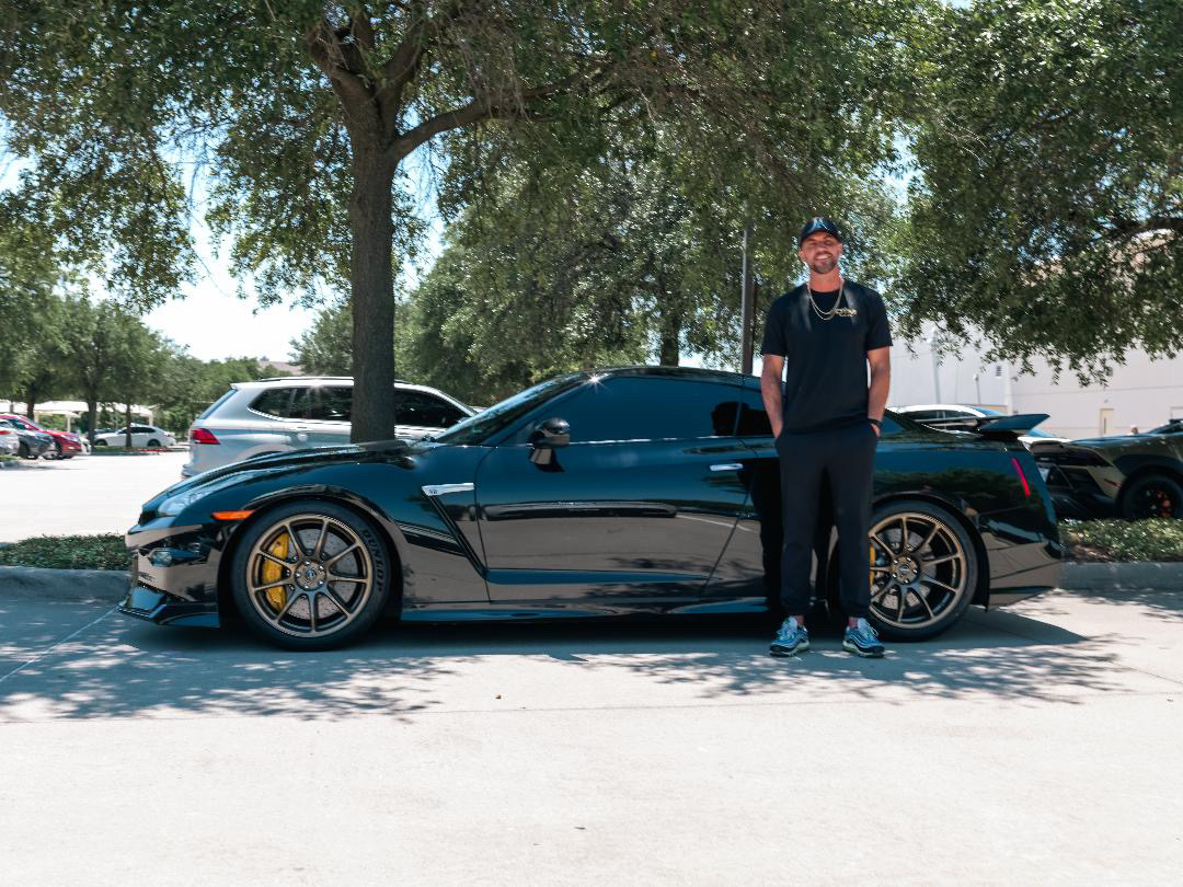 A man stands proudly next to his black Nissan GT-R with aftermarket wheels in a sunny parking lot.