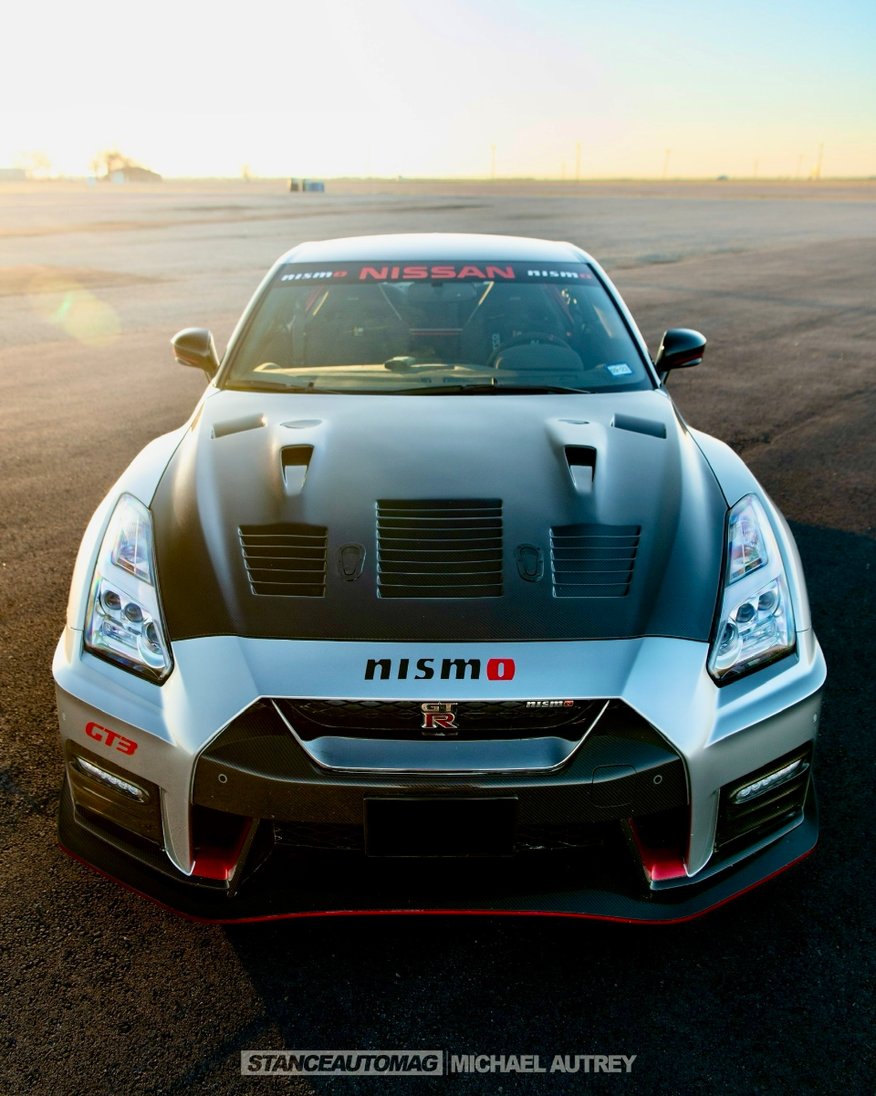 Front view of a silver Nissan GT-R Nismo with a carbon fiber hood and red accents, parked on an airfield at sunset.