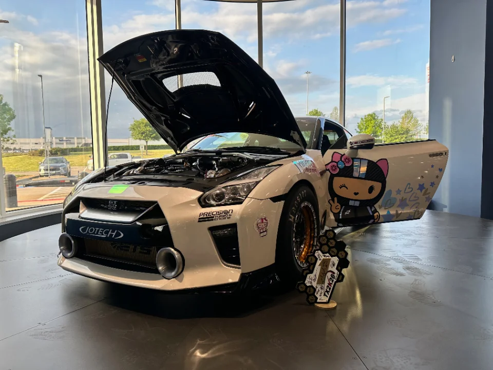 A white Nissan GT-R with custom anime-style livery, open hood, and large intake funnels on display in a showroom with a trophy.
