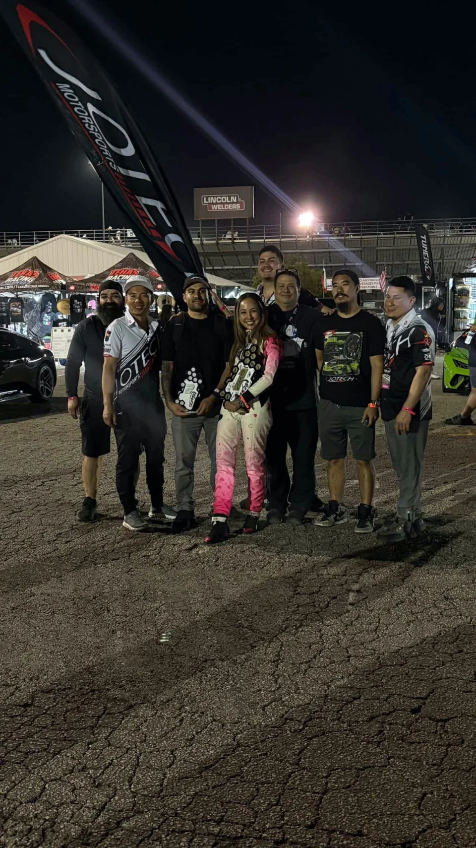A group of Jotech Motorsports team members celebrating with a trophy at a night event, with a Jotech banner in the background.