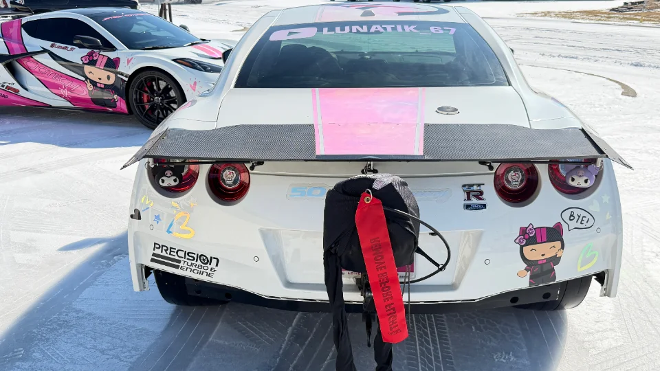 Rear view of a white Nissan GT-R with a carbon fiber wing, parachute, and pink accents on an ice track, with a C8 Corvette in the background.