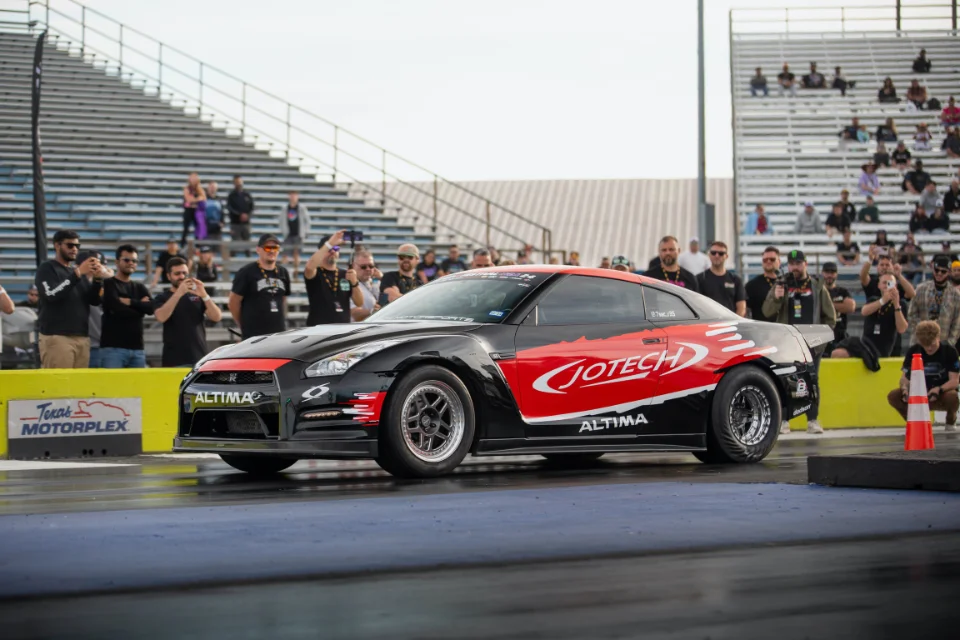 A Jotech Motorsports Nissan GT-R with black and red livery staged on a drag strip, with a crowd of spectators in the stands.