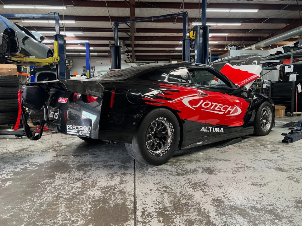 Black Nissan GT-R drag car with red Jotech livery and parachute deployed, parked in a performance tuning shop.