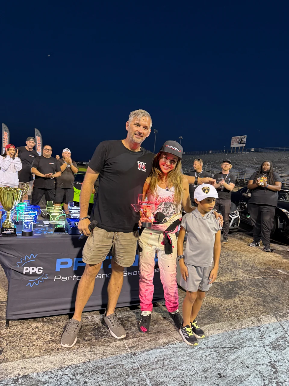 A group of people, including a man, woman, and child, posing with trophies at a drag racing event with a Jotech banner.