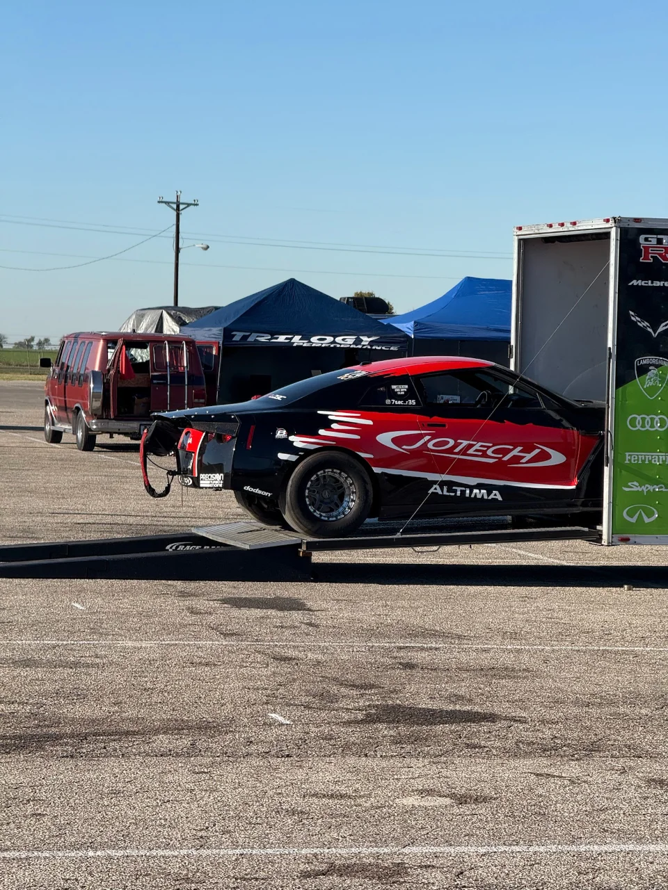 A black and red Jotech Motorsports drag car, possibly an Altima, being unloaded from a trailer at a track event.