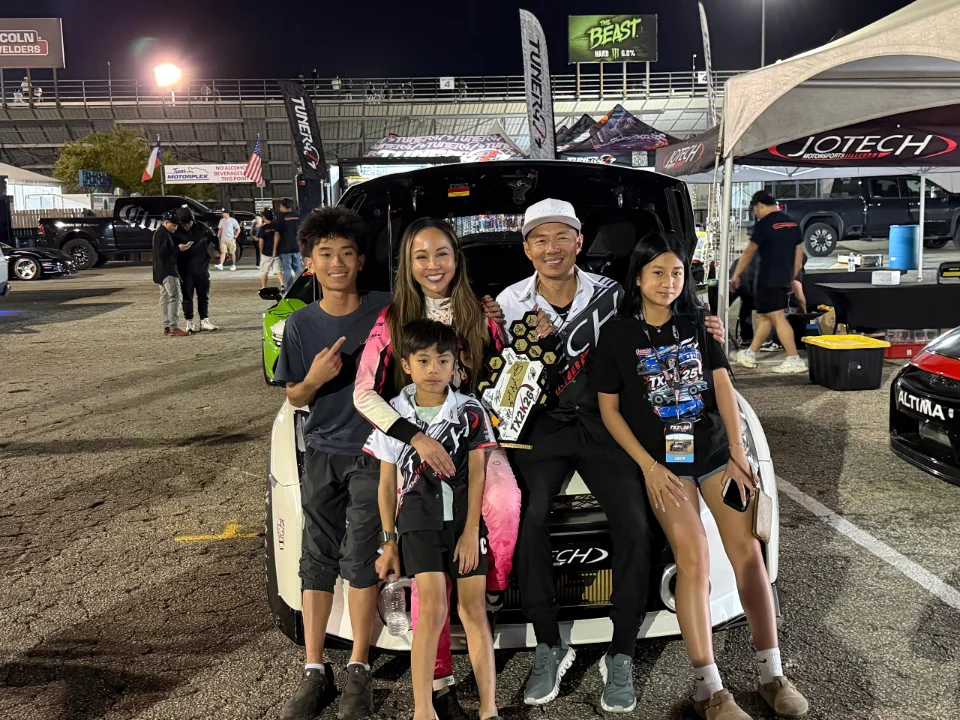 A family poses with a trophy at a night drag racing event, with a Jotech Motorsports tent in the background.