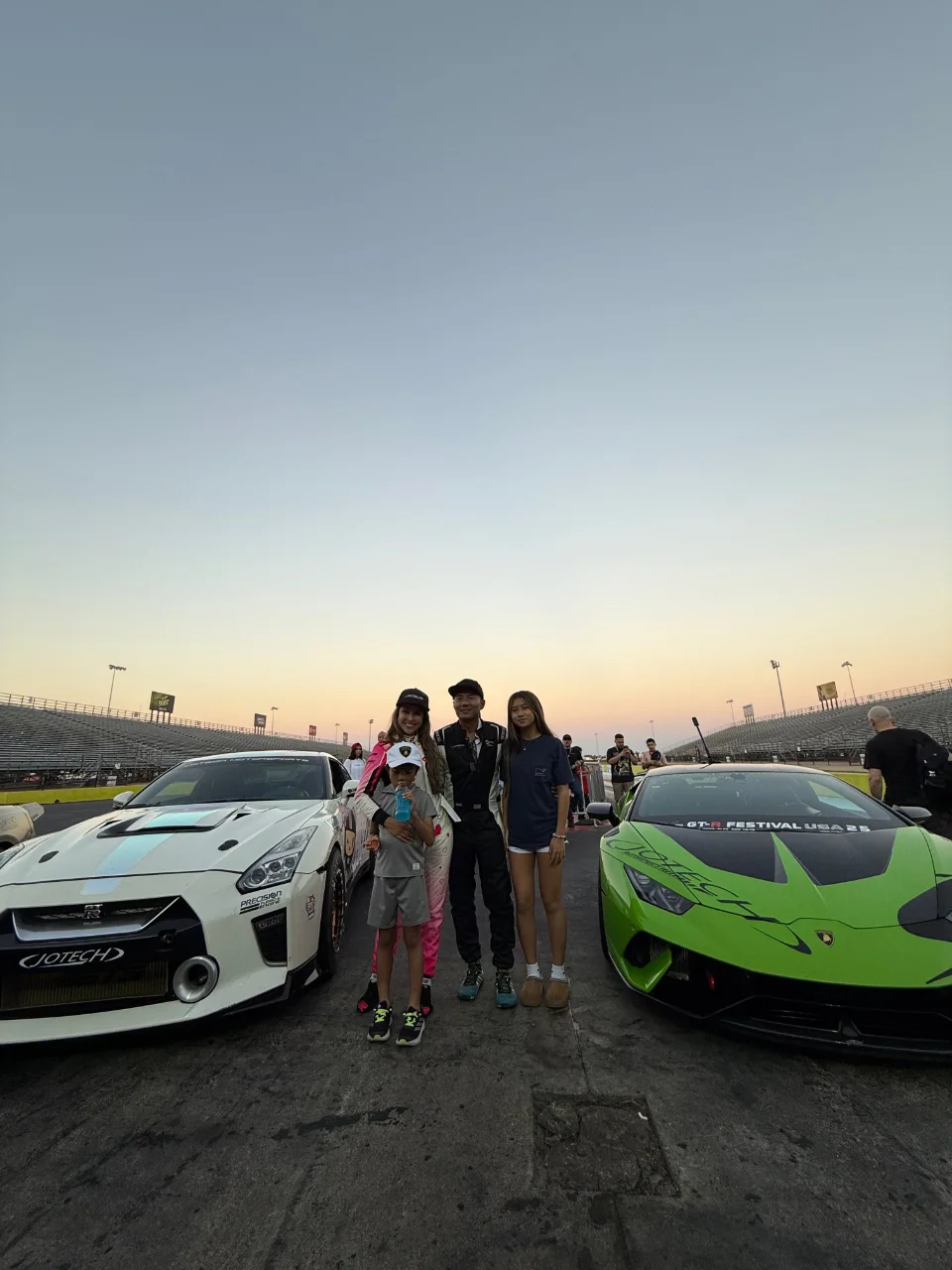 Family and racers posing with a white Nissan GT-R and a green Lamborghini Huracan at a drag strip.