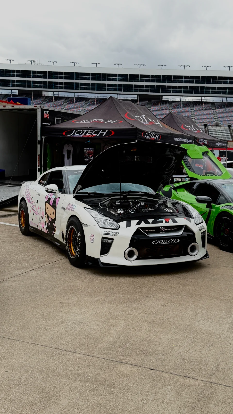 A white Nissan GT-R with its hood open, featuring custom livery, parked next to a green Lamborghini under Jotech Motorsports tents at a track event.