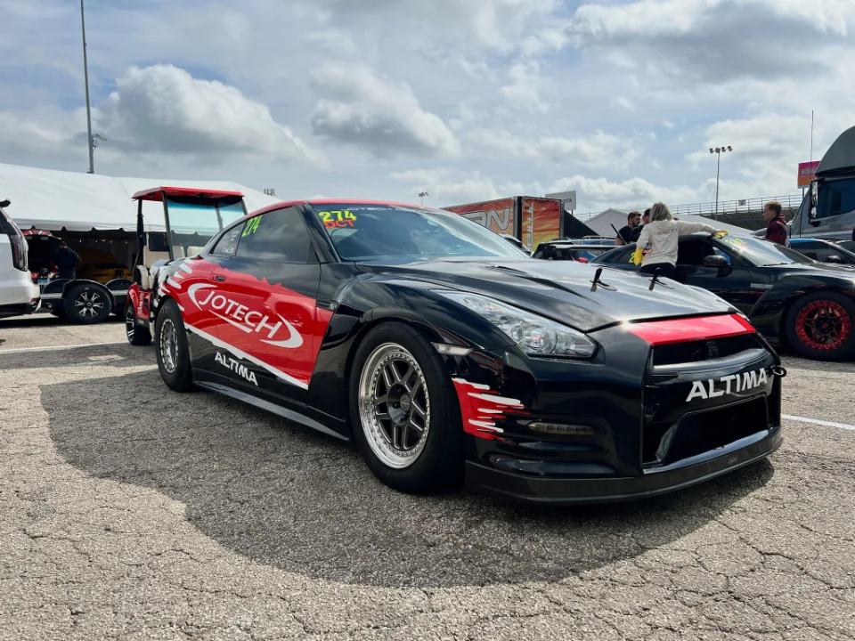 A black and red Jotech Nissan GT-R drag car with racing livery parked at a track, with tents and other vehicles in the background.