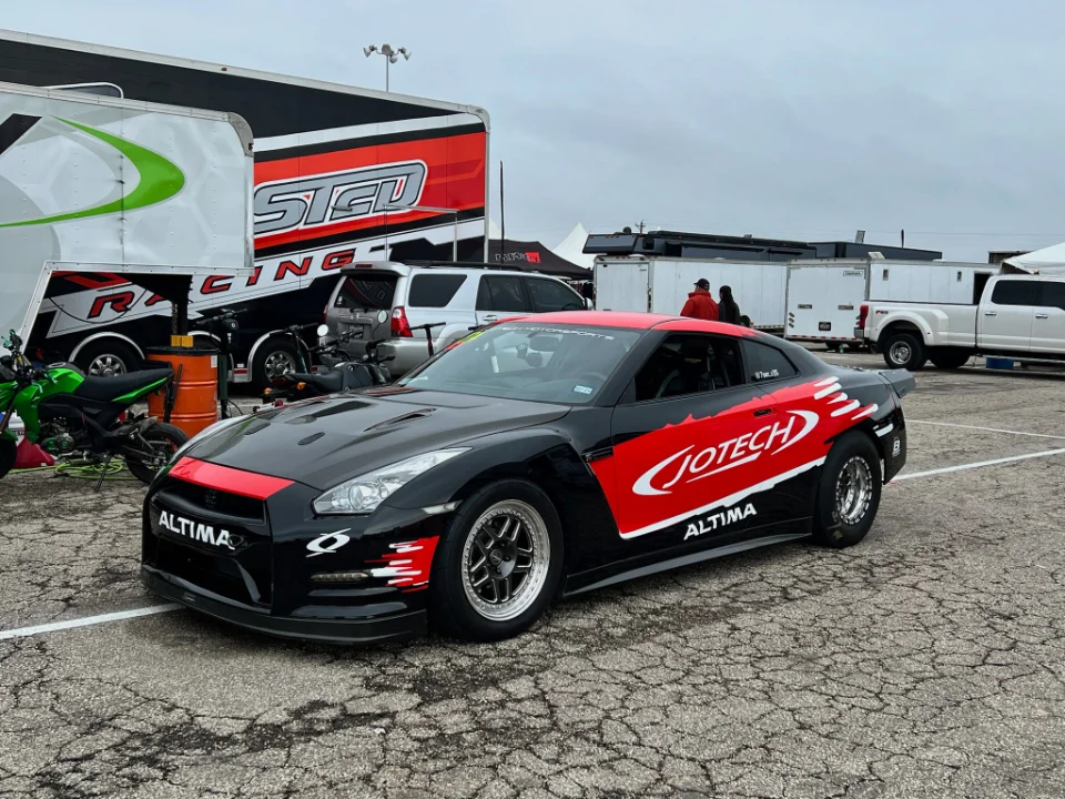 A black and red Jotech Motorsports Nissan GT-R drag car parked at a race track paddock with trailers in the background.