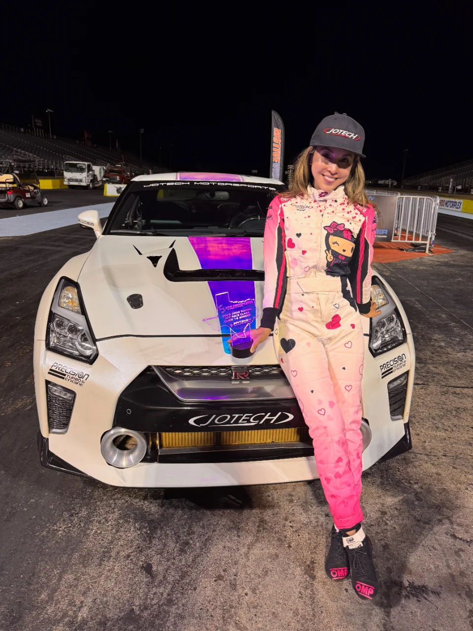A woman in a pink racing suit stands next to a white Nissan GT-R race car with Jotech Motorsports livery at a drag strip.
