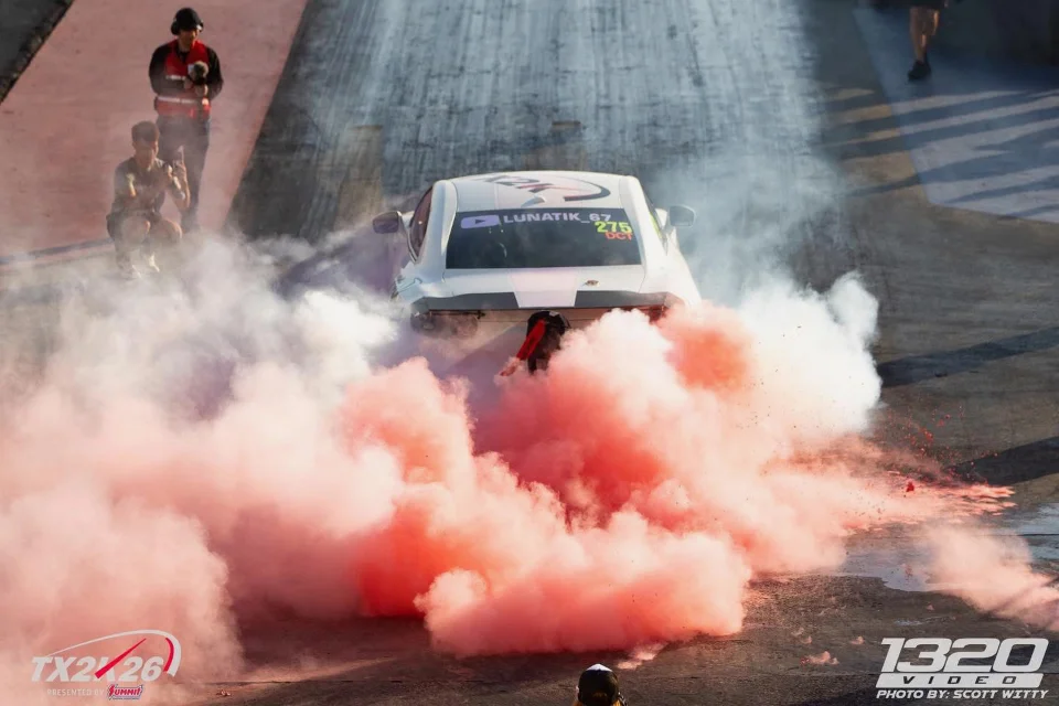 White drag car performing a burnout with vibrant red smoke on a drag strip, with spectators in the background.