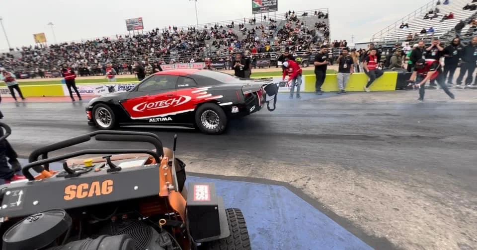 A Jotech Motorsports Nissan Altima drag car performing a burnout on a drag strip with a crowd in the stands.