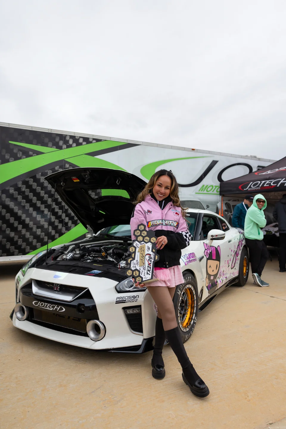 A woman in a pink jacket holding a trophy stands proudly next to a white Nissan GT-R race car with custom livery at a Jotech Motorsports event.