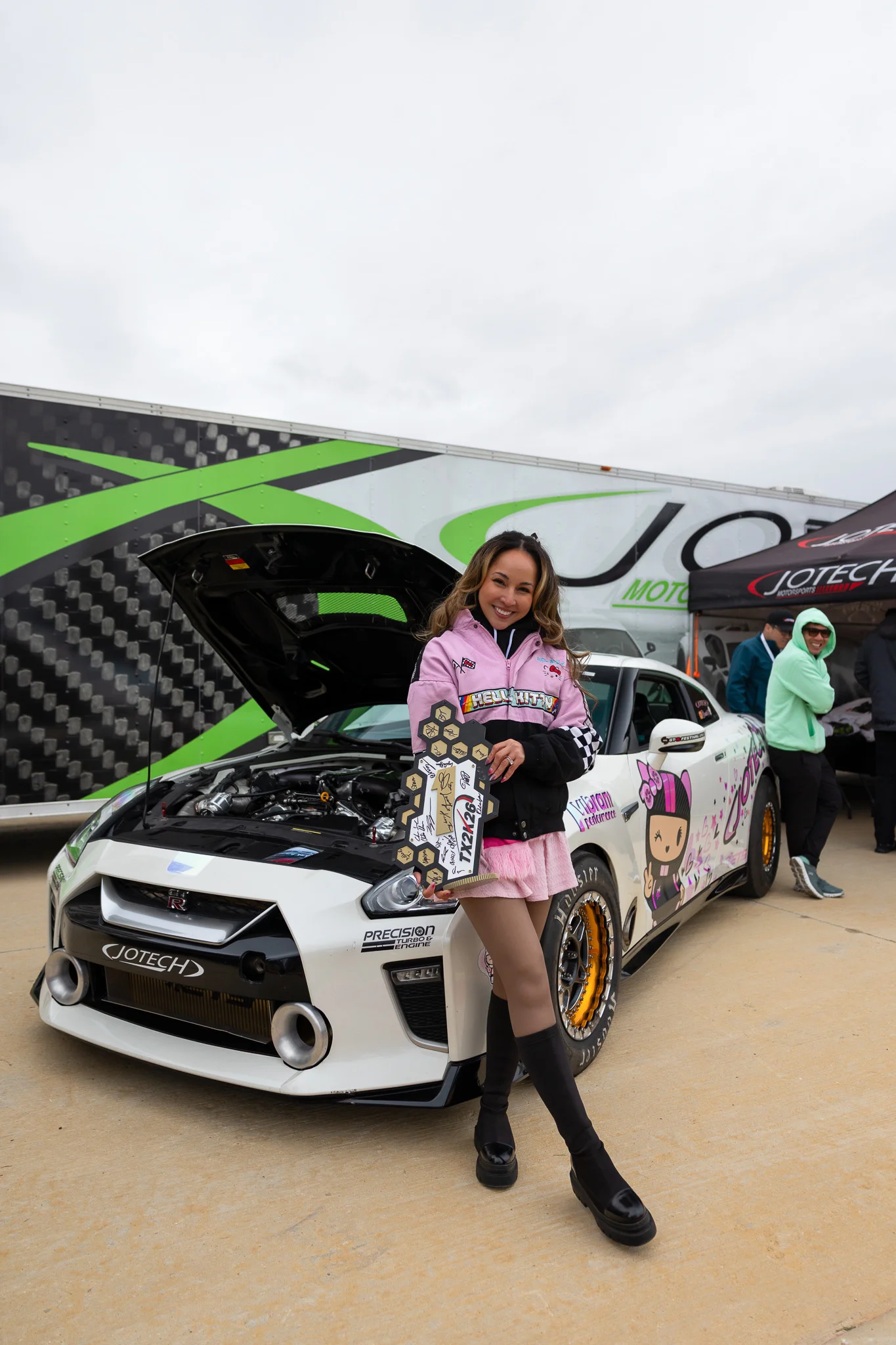 A woman in a pink jacket holding a trophy stands proudly next to a white Nissan GT-R race car with custom livery at a Jotech Motorsports event.