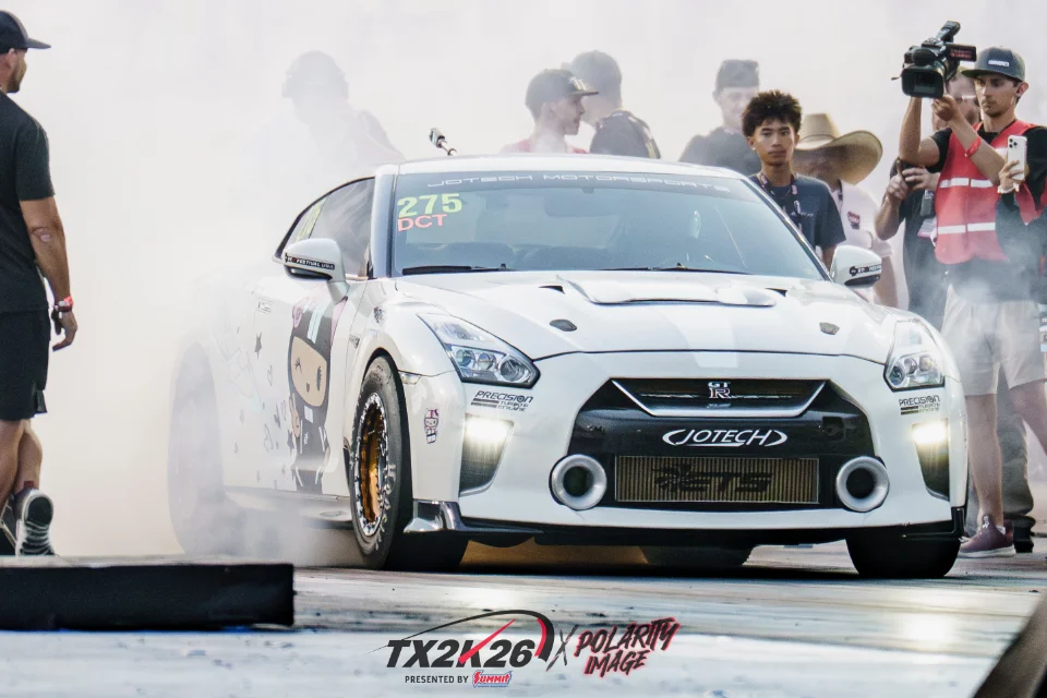 White Nissan GT-R performing a burnout on a drag strip at TX2K, with Jotech Motorsports branding and spectators.