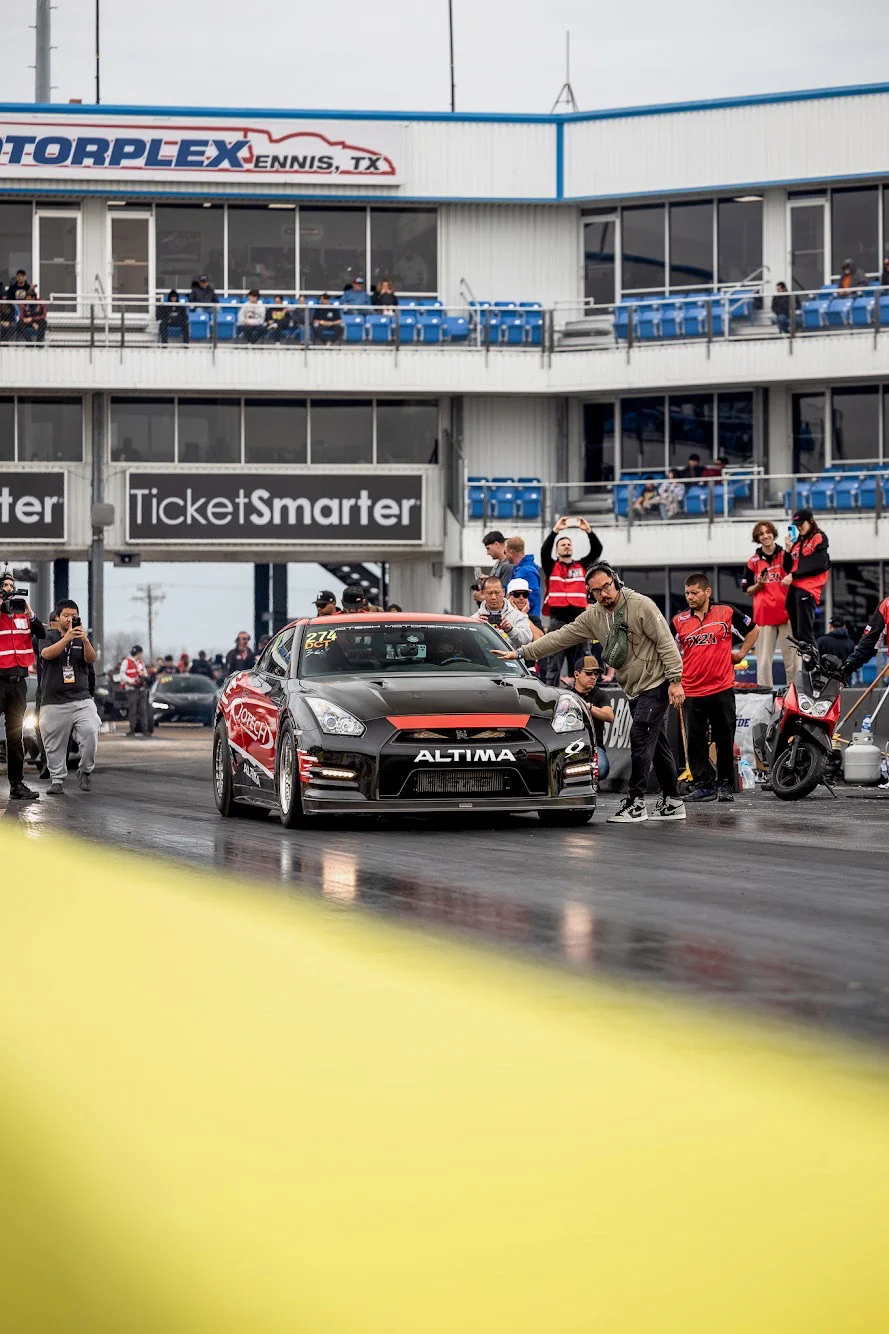 A black Nissan GT-R with red accents and Jotech livery at the starting line of a drag strip, with spectators and a timing tower.