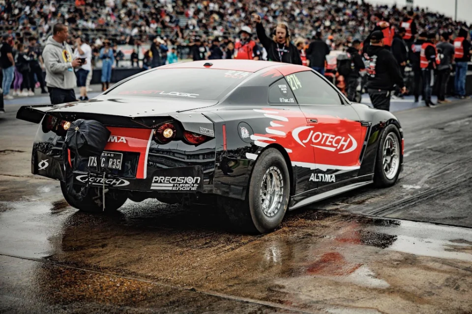 A black and red Jotech Nissan GT-R drag car with a parachute at a wet drag strip, surrounded by spectators.