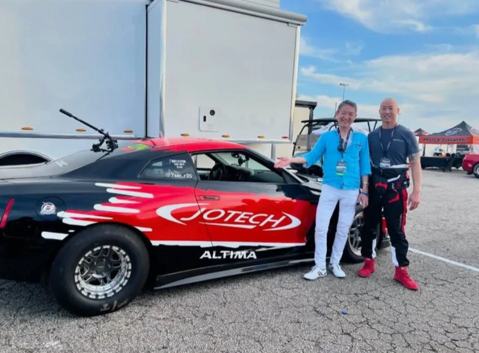 Two men pose next to a black and red Jotech Nissan Altima drag car with racing slicks at a track event.