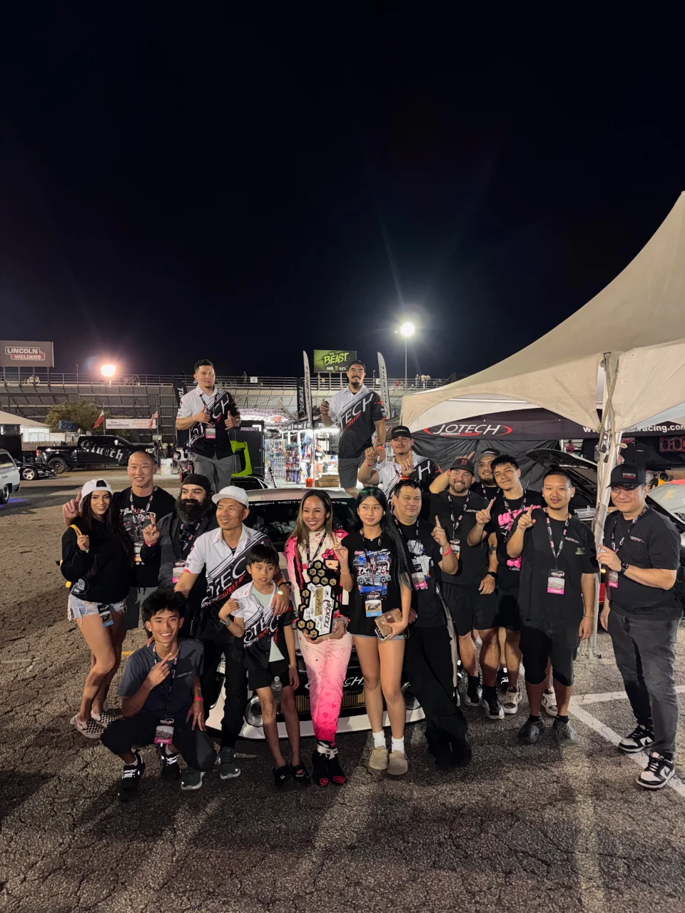 Jotech Motorsports team and supporters celebrate with trophies at a nighttime drag racing event, in front of a car and tents.