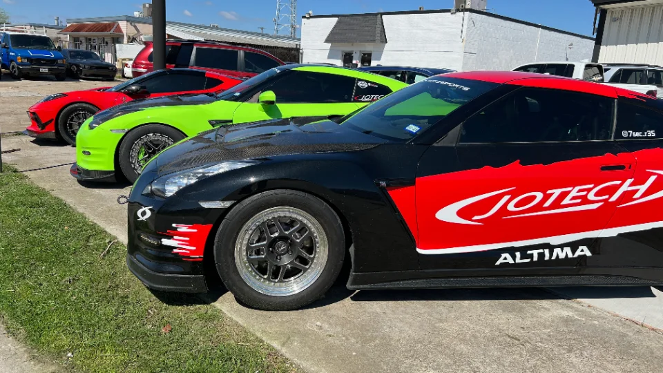 A black and red Jotech Nissan GT-R, a green Nissan GT-R, and a red Corvette parked outside a performance shop.