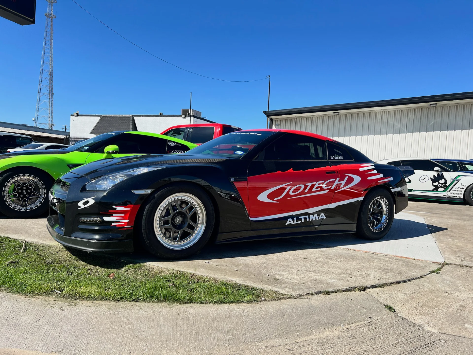 Black Nissan GT-R with Jotech livery and a green Chevrolet Camaro parked outside a performance tuning shop on a sunny day.