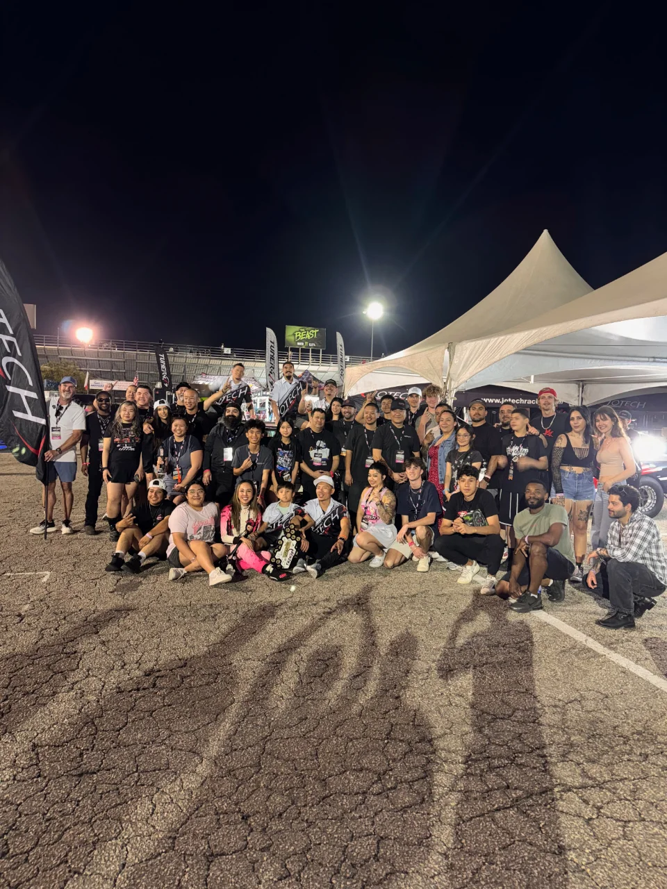 A large group photo of the Jotech Motorsports team and supporters gathered at a night event, with tents and banners in the background.
