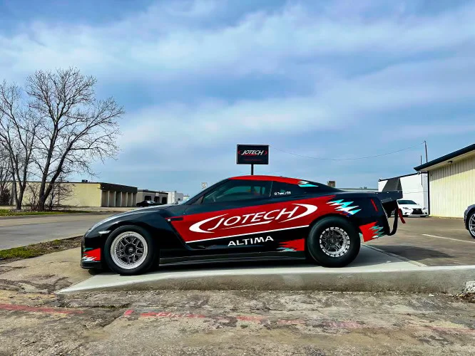 A Jotech Motorsports Nissan GT-R drag car with custom red and black livery and drag wheels parked outside a shop.