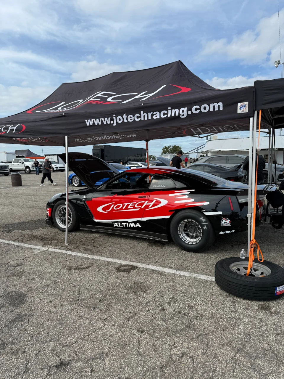 A black and red Jotech Racing Nissan GT-R drag car parked under a Jotech tent at a track event, with other cars in the background.