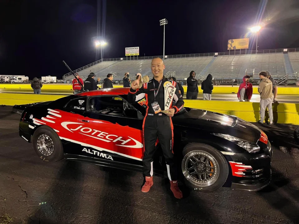 A driver holding a trophy next to a black and red Jotech Nissan GT-R drag car at a nighttime drag strip.
