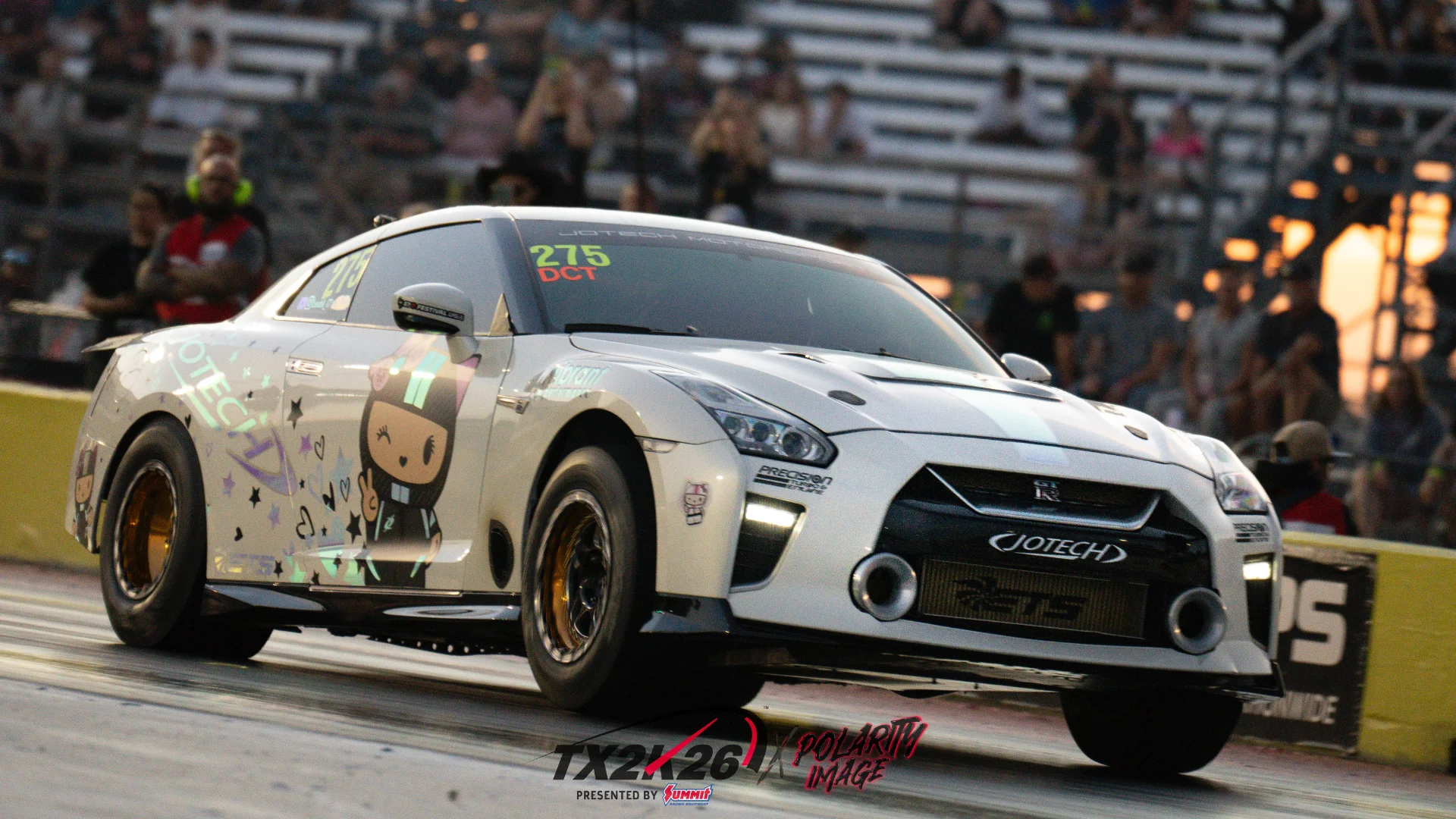 A white Nissan GT-R race car with Jotech branding launches on a drag strip, with spectators in the background at sunset.