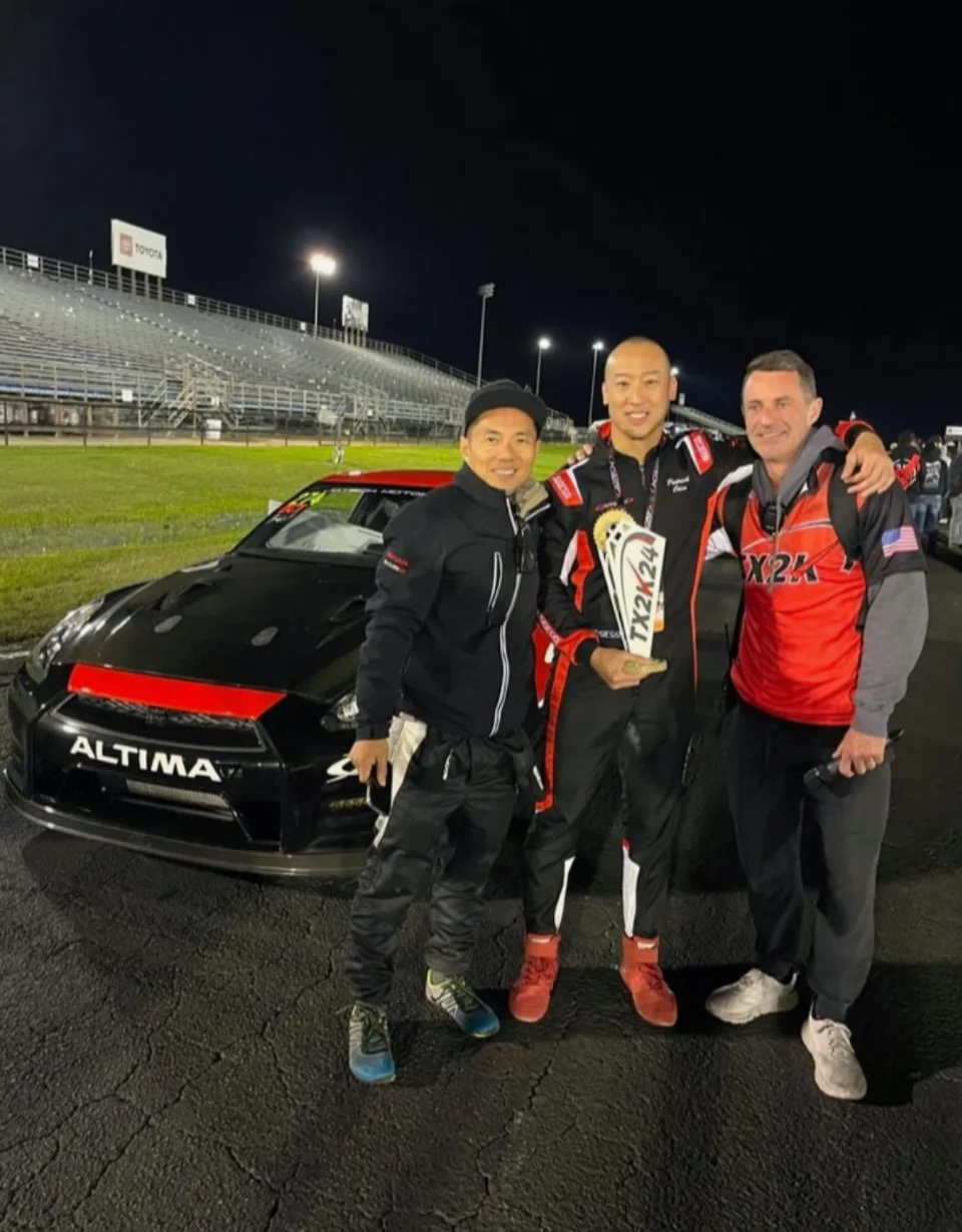 Three men celebrate with a trophy in front of a black Nissan GT-R at a drag strip at night.