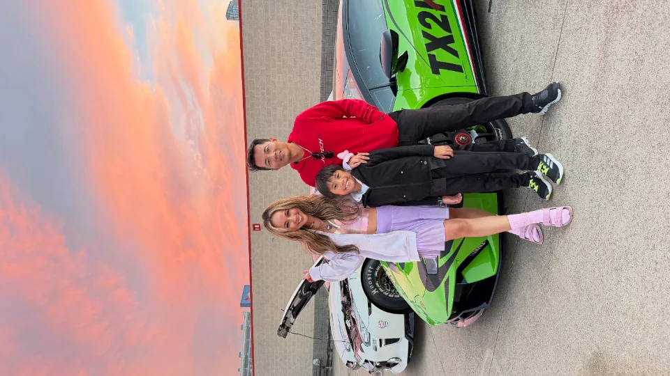 A family poses with a green Lamborghini Huracan race car with TX2K livery at a track event during sunset.