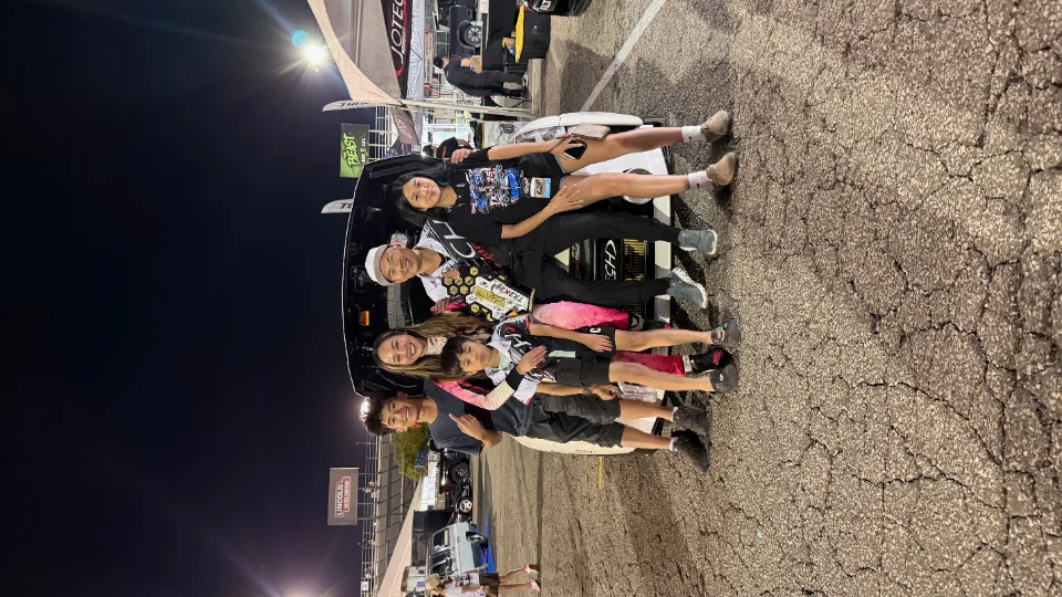 A family poses with trophies in the trunk of a white car at a night drag racing event.