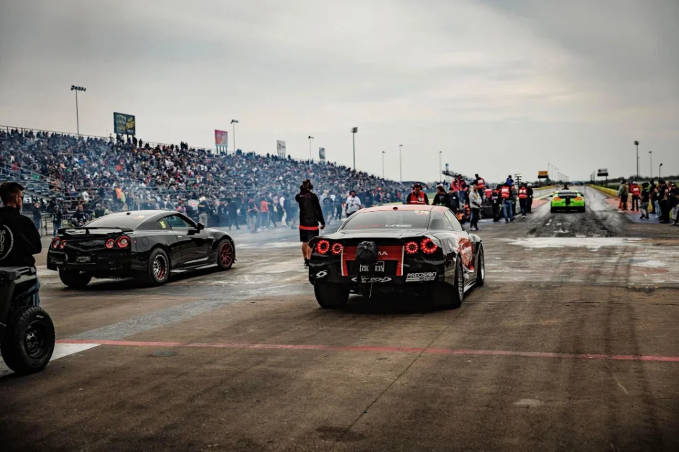 Two Nissan GT-Rs at a drag strip, one performing a burnout, with a crowd in the background.