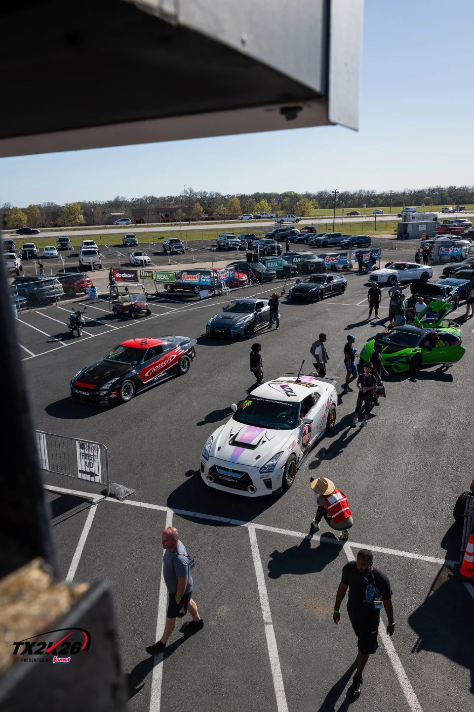 Overhead view of multiple custom Nissan GT-Rs and other performance cars parked in a pit area at a track event, with people walking around.