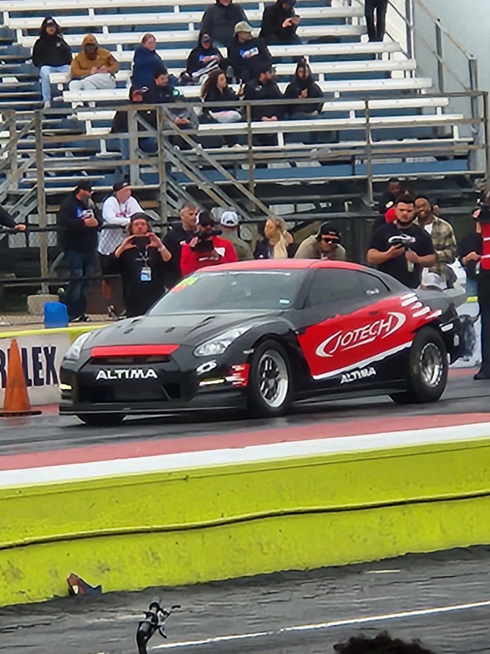 A black and red Jotech Motorsports Nissan GT-R at the starting line of a drag strip with spectators in the stands.