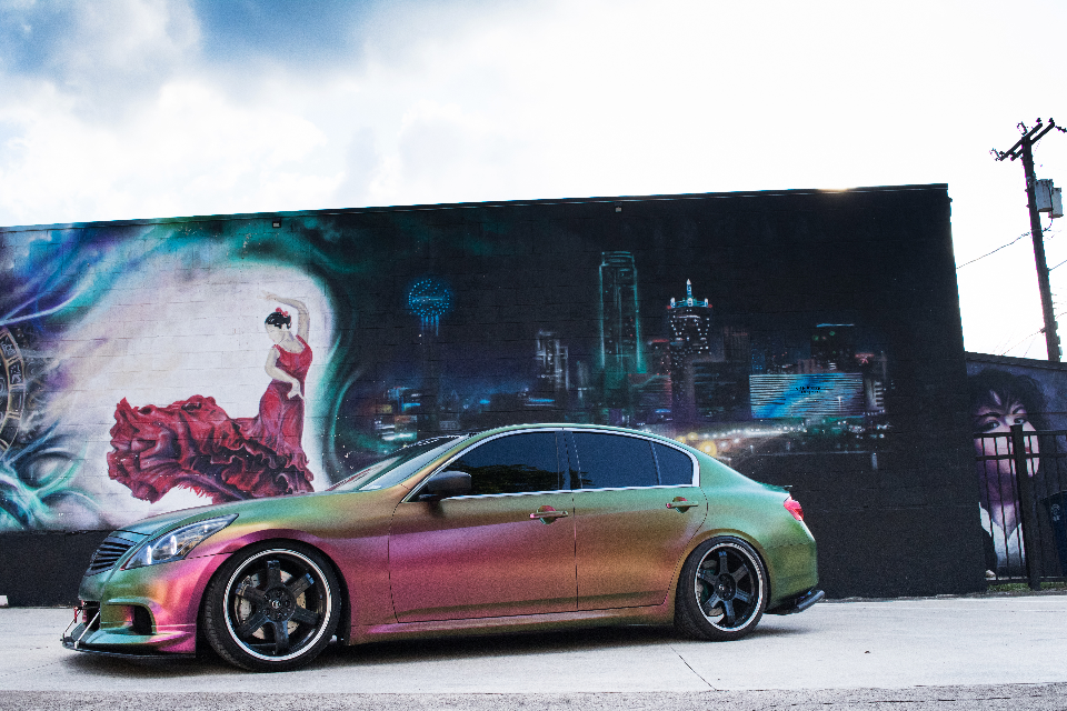 A chameleon-wrapped Infiniti G37 sedan parked in front of a vibrant street art mural featuring a dancer and city skyline.