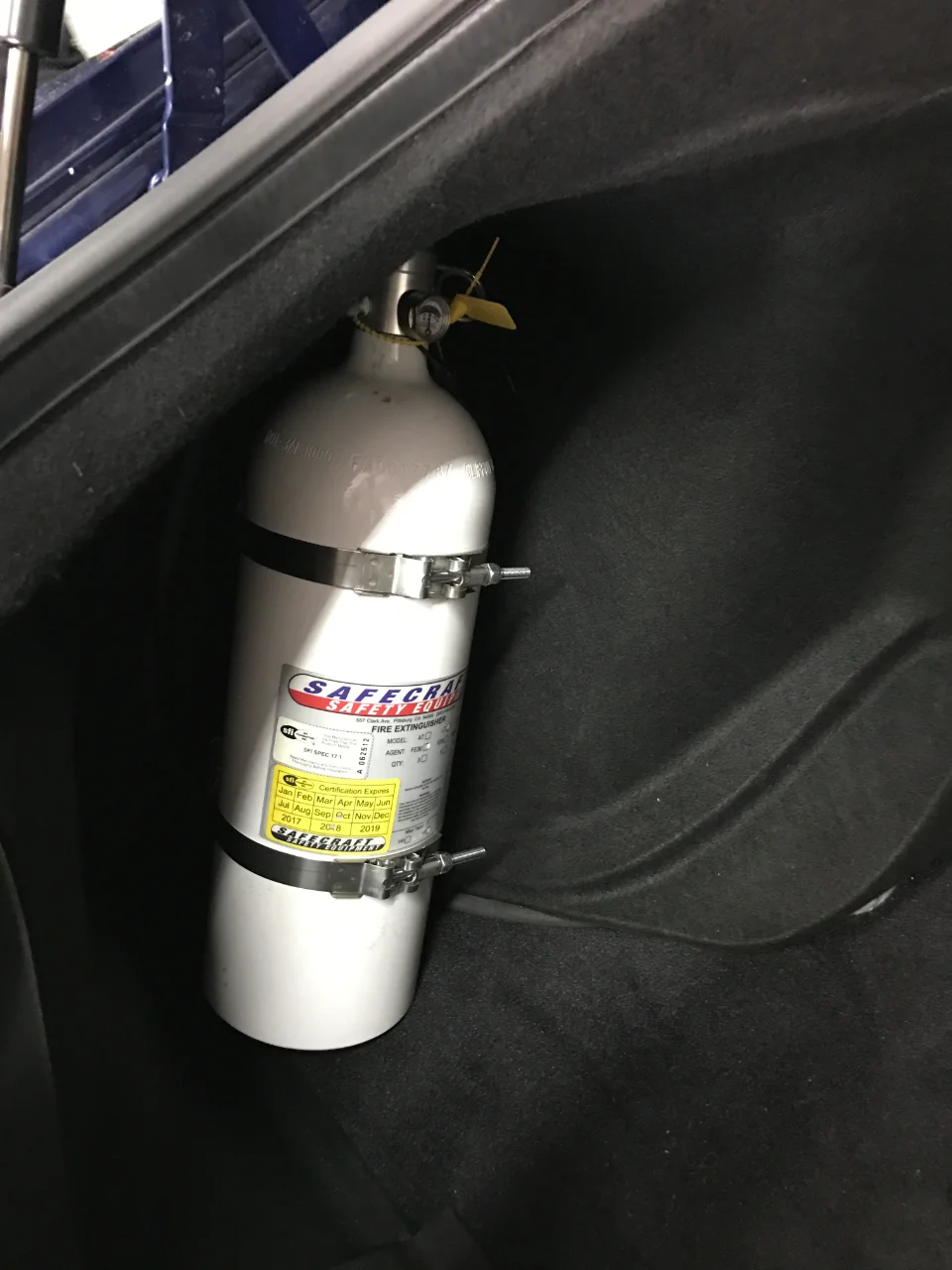 A Safecraft fire extinguisher mounted in the trunk of a car, secured with metal straps.