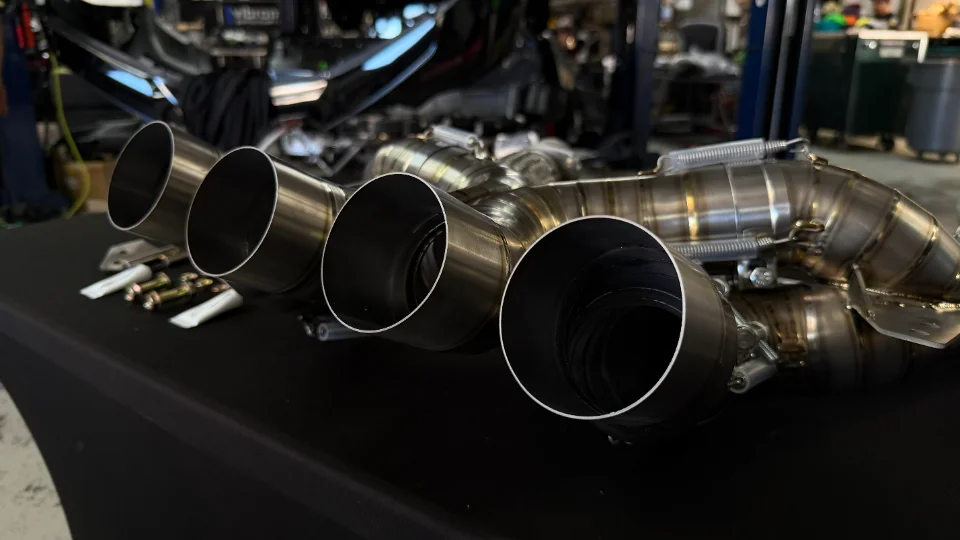 Close-up of a new, polished aftermarket exhaust system with multiple tips and mounting hardware on a table in a shop.