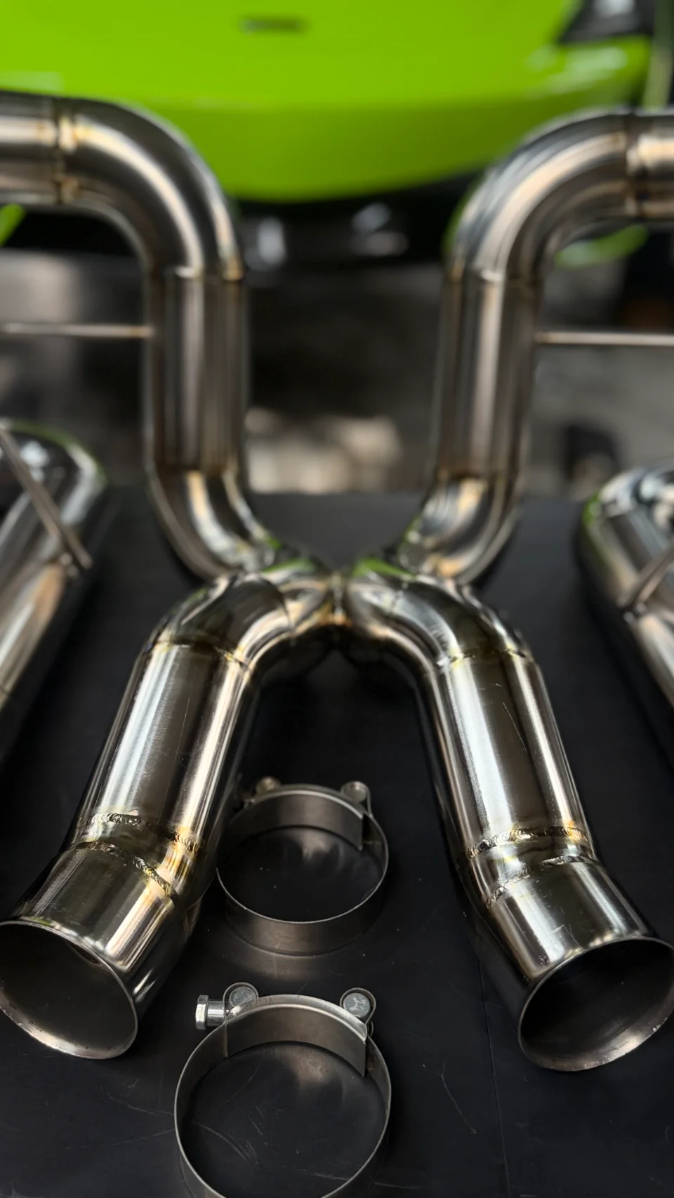 A close-up of a polished stainless steel aftermarket exhaust X-pipe with clamps, with a blurred green Lamborghini in the background.