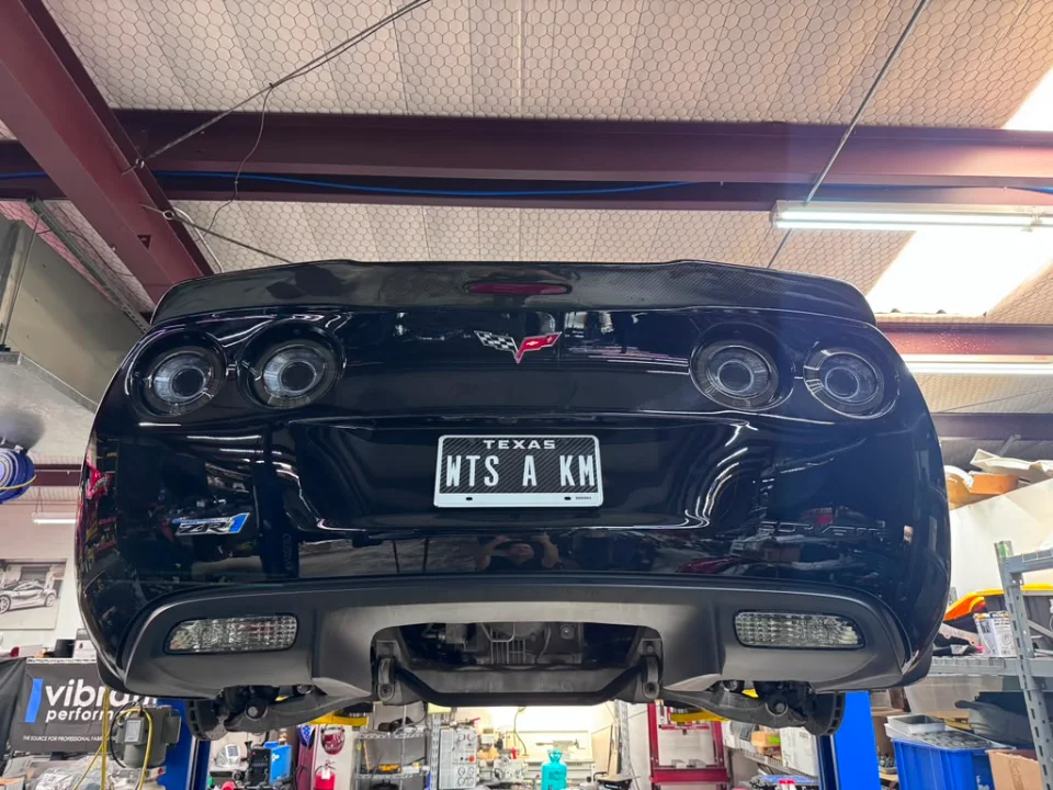 Rear view of a black Corvette ZR1 on a lift in a performance tuning shop, featuring a carbon fiber spoiler and Texas license plate.